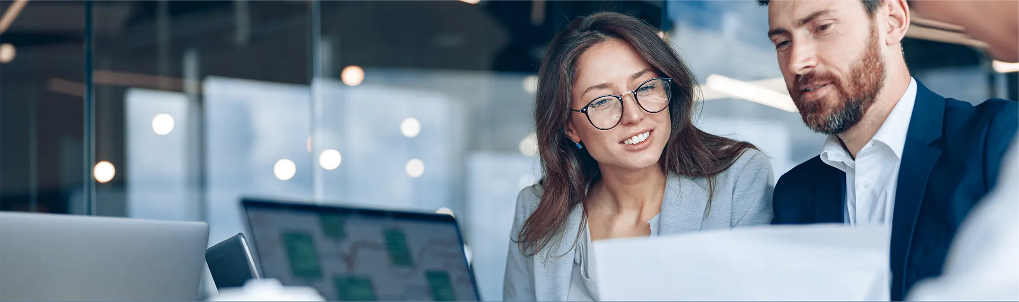 Smiling business woman wearing glasses and male colleague collaborating on laptop computer in contemporary office workspace environment