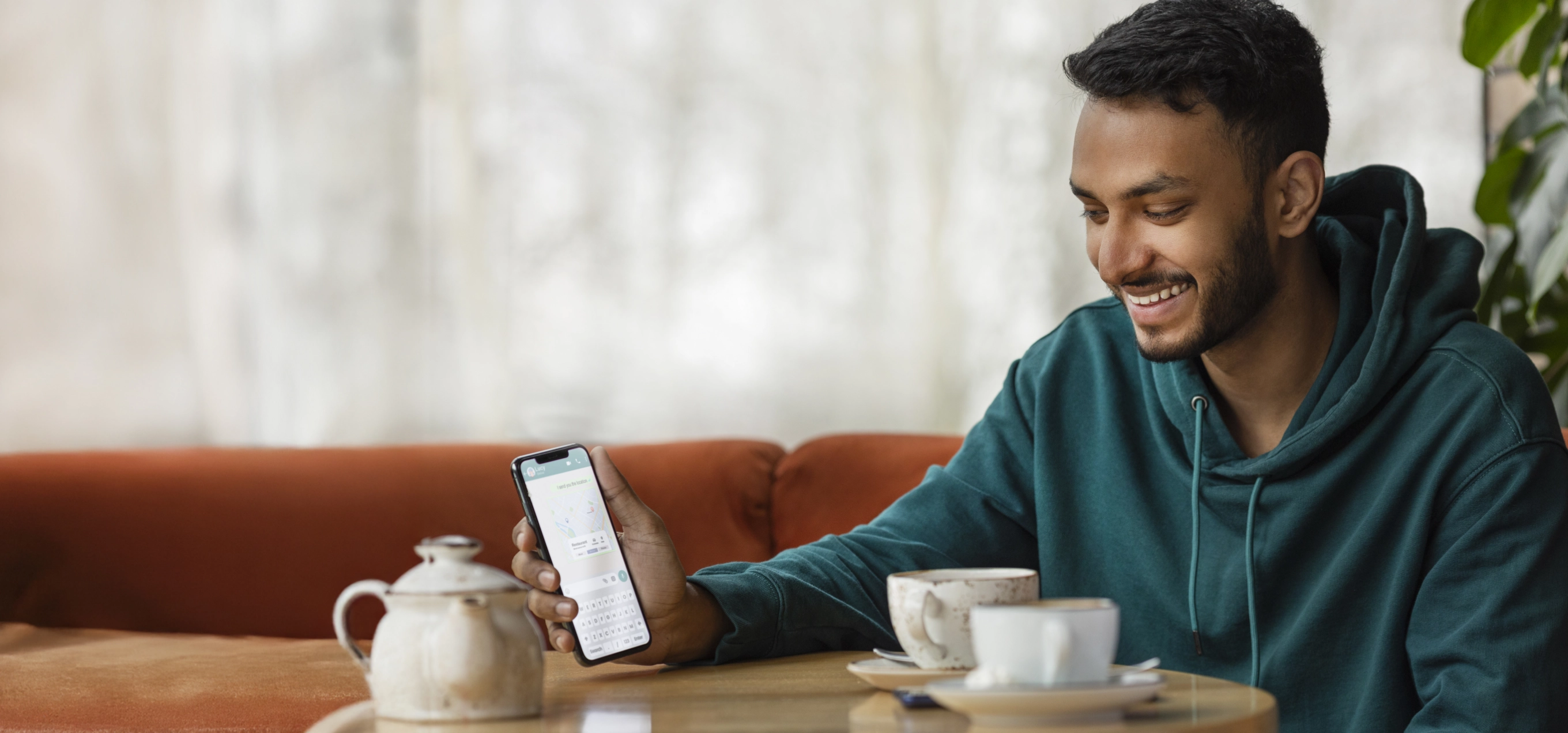 Smiling young man in green hoodie using smartphone at cafe table with coffee cup and teapot in cozy interior setting