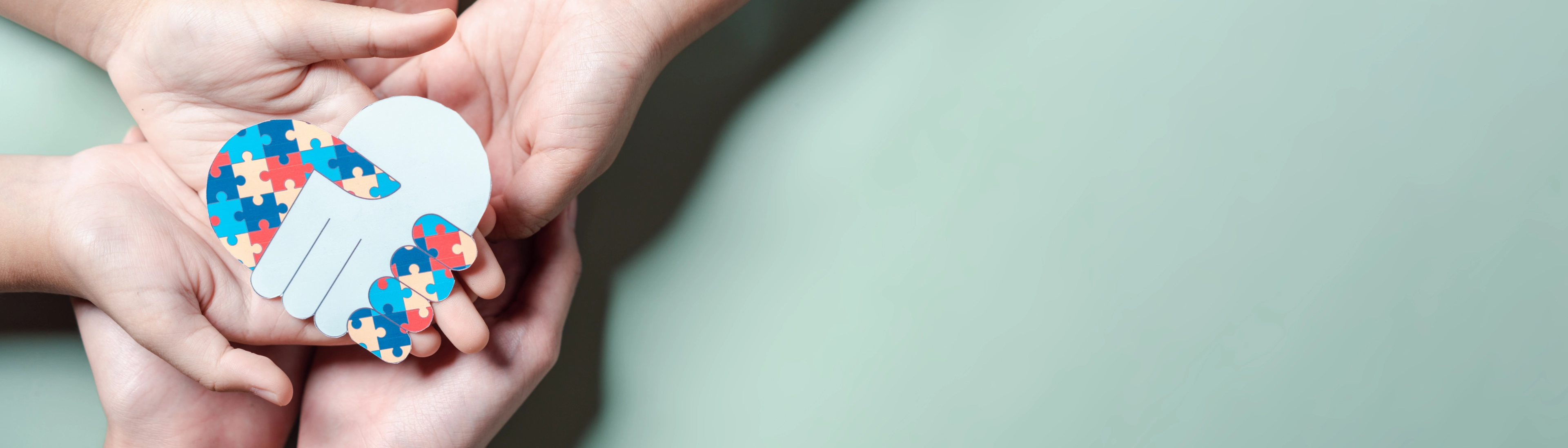 Hands cradling paper brain with colorful puzzle pieces on mint background representing autism awareness and mental health