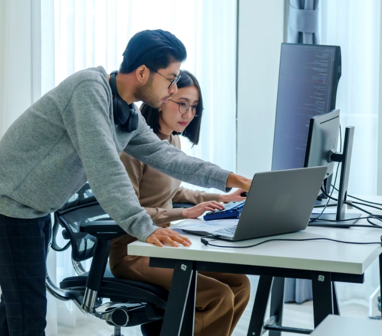Two software developers collaborating on laptop in modern office with whiteboard and sticky notes representing teamwork
