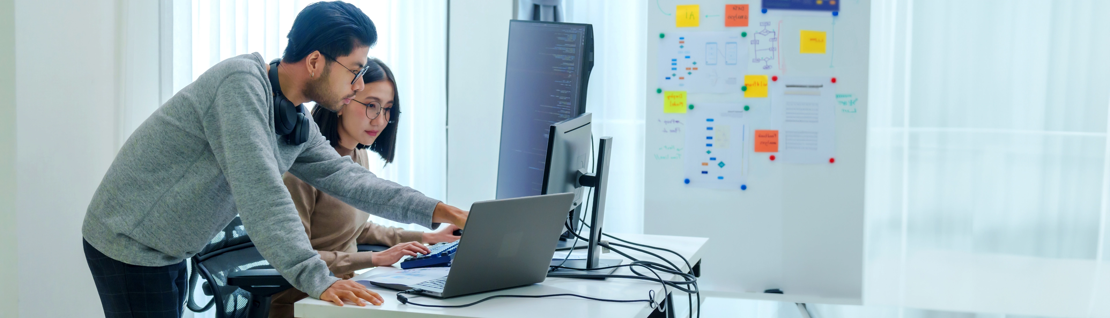 Two software developers collaborating on laptop in modern office with whiteboard and sticky notes representing teamwork